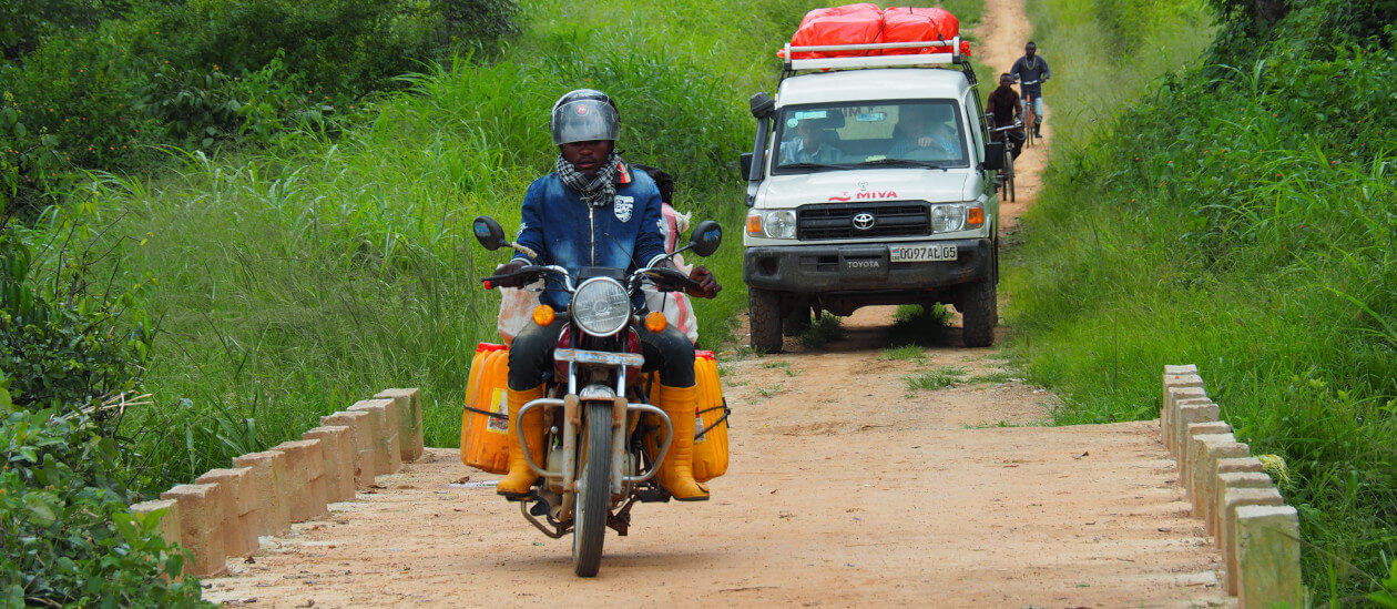 A person rides over a dirt bridge on a motorbike loaded with yellow containers, followed by a white SUV with MIVA lettering and red cargo on the roof, and travels through a lush, green rural area