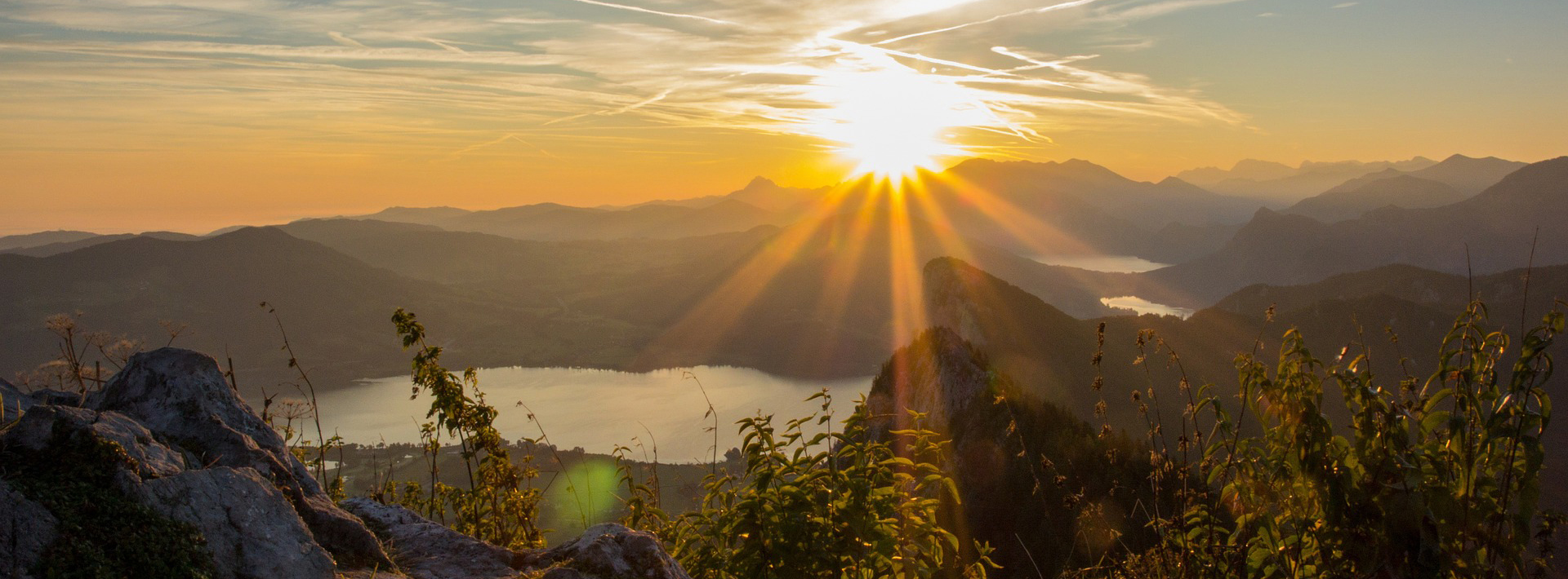 Die Sonne geht über den Bergen unter und taucht eine malerische Landschaft mit einem See, Hügeln und Vegetation im Vordergrund in goldenes Licht. 