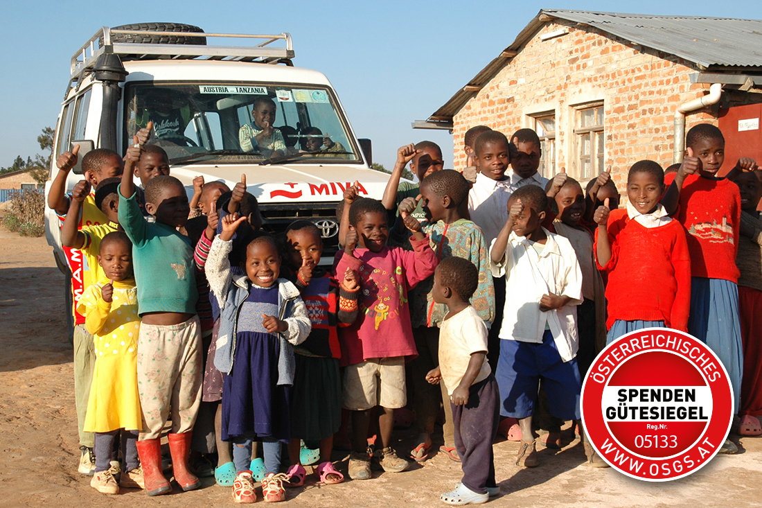 A group of smiling children stand in front of a brick building and raise their hands. Behind them is a white vehicle with the word ‘MIVA’ on the front. A superimposed text and the red Austrian donation seal of approval indicate that donations are tax-deductible.