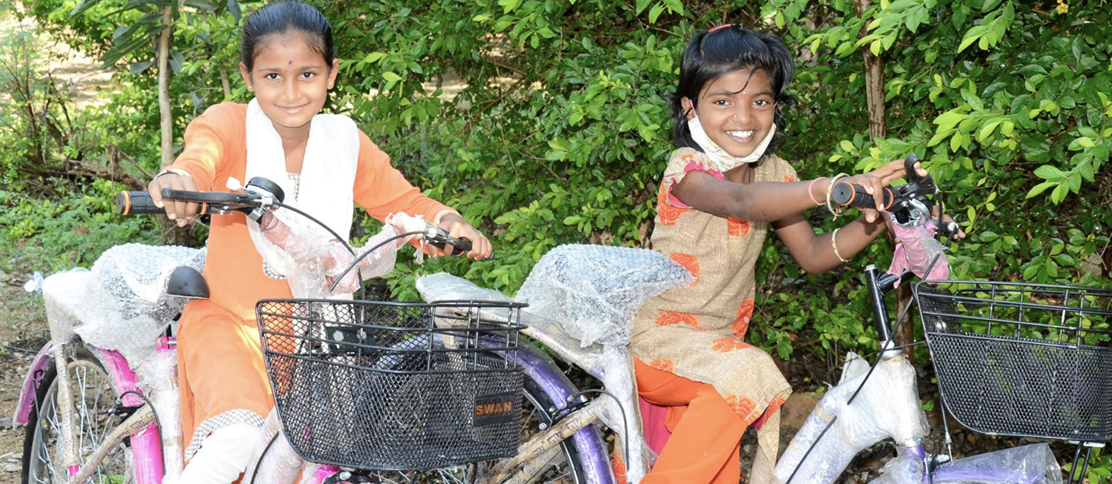 Two young girls smile on their new, purple bicycles outside. The bikes have baskets and are still partially wrapped in plastic film.