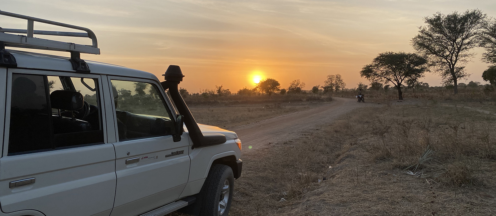 Ein weißes Fahrzeug parkt bei Sonnenuntergang auf einer unbefestigten Straße in einer trockenen, grasbewachsenen Landschaft.