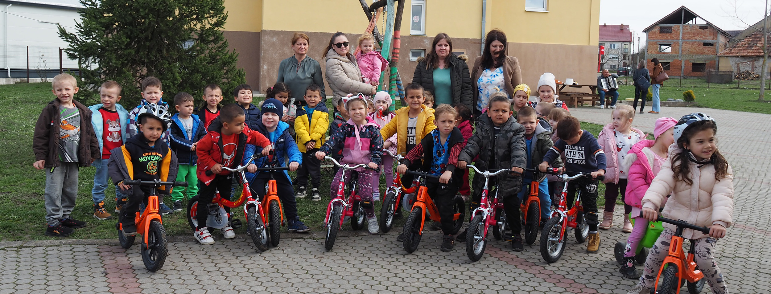 Eine Gruppe junger Menschen, teils stehend, teils sitzend auf MIVA-Fahrrädern, posiert im Freien, hinter ihnen mehrere Erwachsene in der Nähe eines gelben Gebäudes. Die Kinder wirken fröhlich.