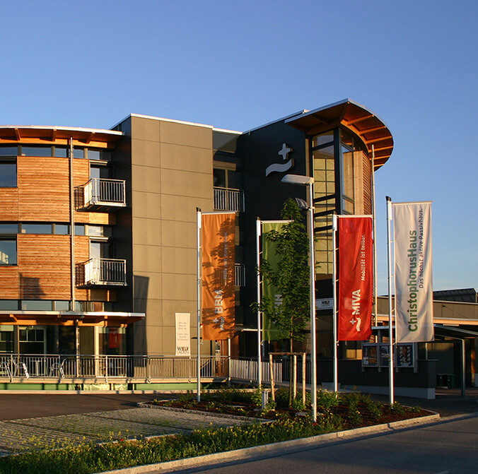 The ChristophorusHaus, a modern building, combines wood and grey panels in the façade. Large windows break through the front, while four colourful flags wave in front of it in the sunlight.