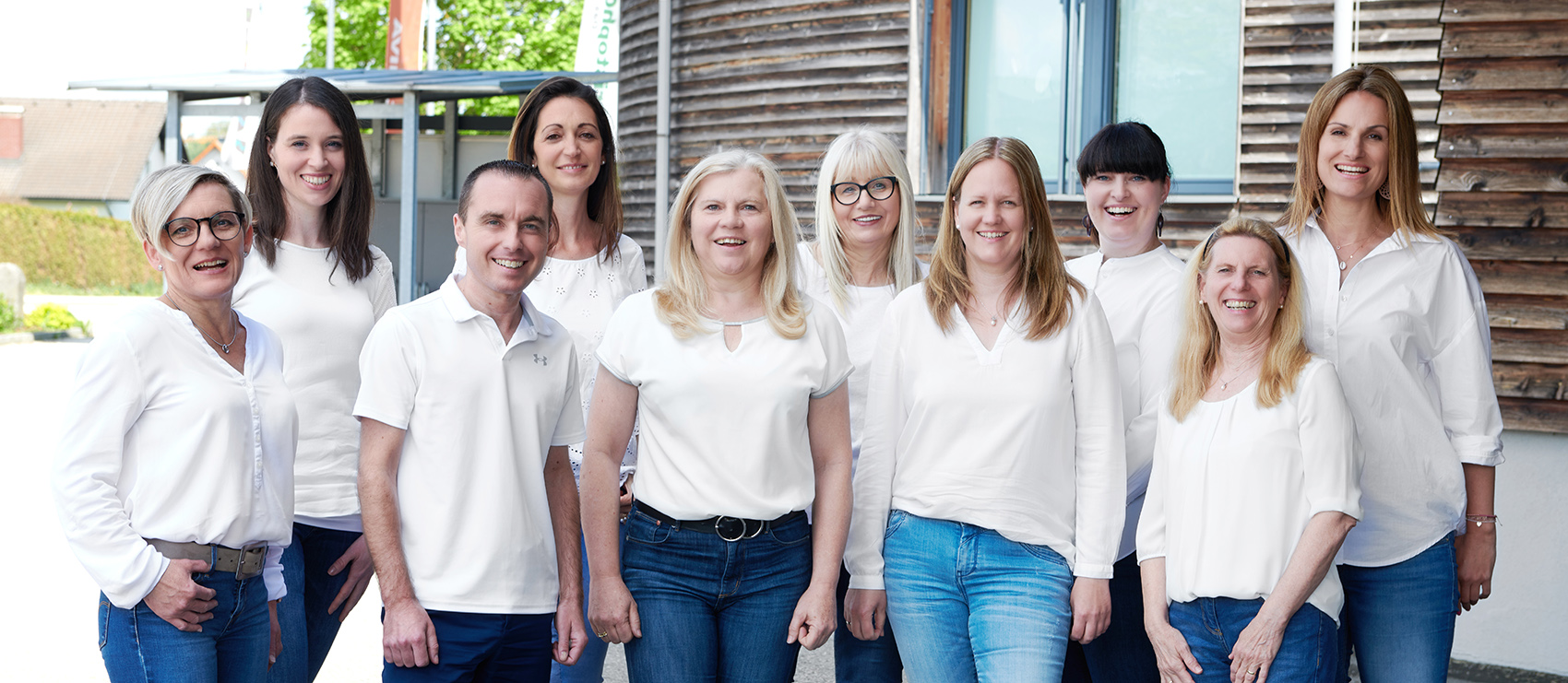 The Miva team, a group of ten people, stand together outside a wooden building, all smiling and wearing white tops with jeans