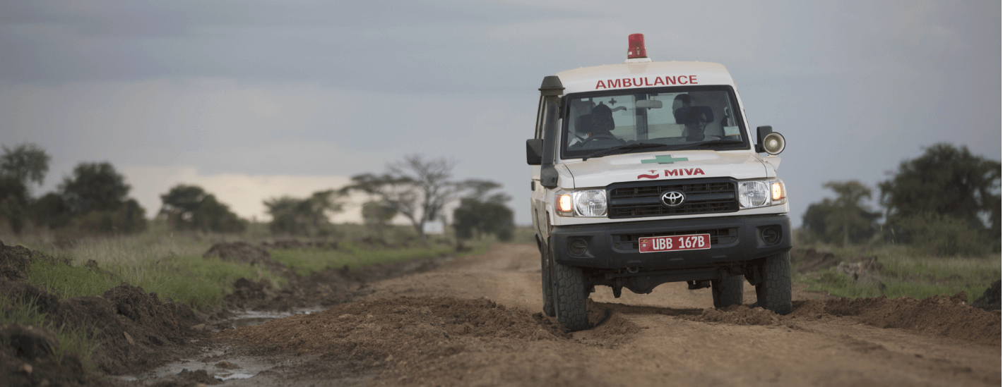 A white Toyota MIVA ambulance with red lights on the roof drives along a muddy, unpaved country road, providing medical care to remote areas surrounded by grass and trees under a cloudy sky.