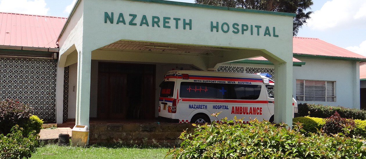 Ein weißer Krankenwagen mit der Aufschrift „Nazareth Hospital“ steht unter dem Vordach eines Gebäudes mit einem Schild mit der Aufschrift „Nazareth Hospital“. Das Gebäude hat ein rotes Dach und ist von grünen Büschen umgeben.
