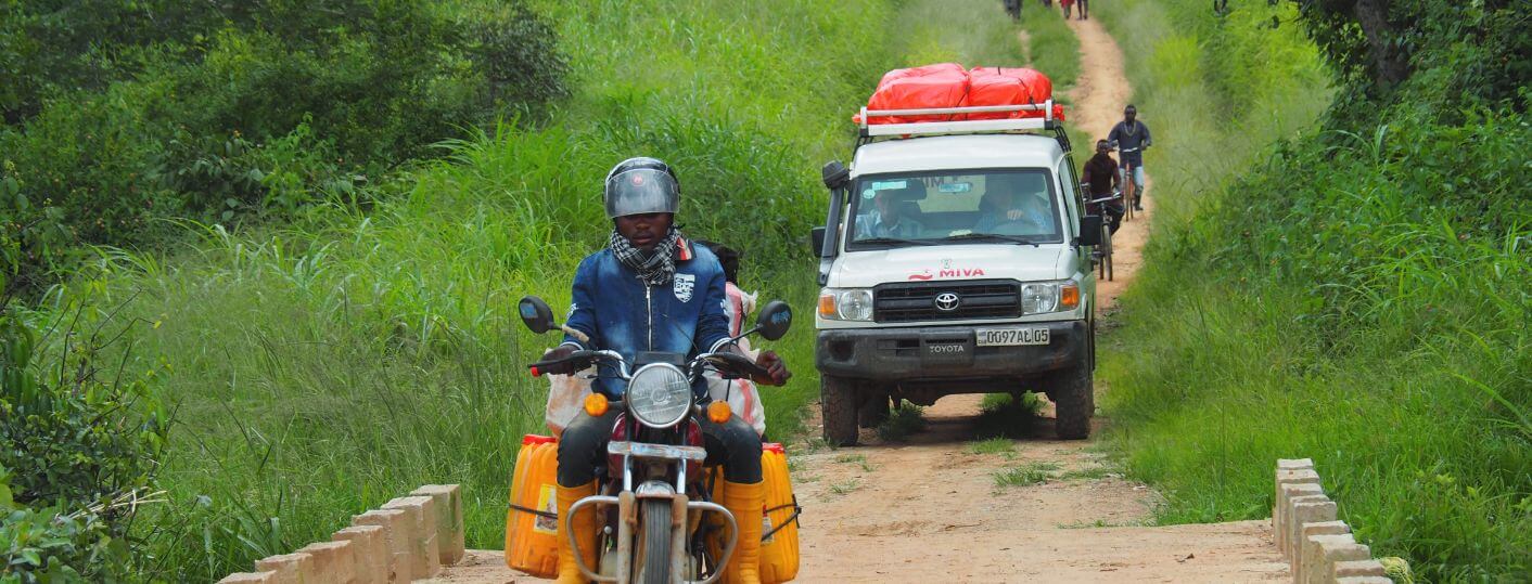 A person rides over a dirt bridge on a motorbike loaded with yellow containers, followed by a white SUV with MIVA lettering and red cargo on the roof, and travels through a lush, green rural area