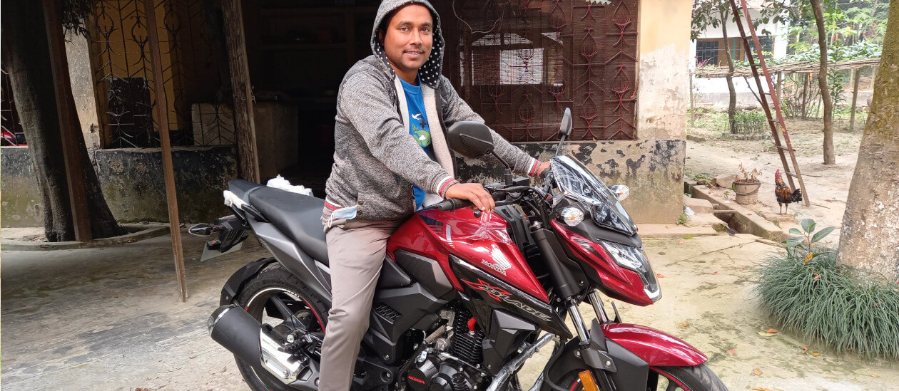 Ein Mann in grauem Kapuzenpullover und beiger Hose sitzt auf einem roten Honda  Motorrad und lächelt in die Kamera. 
